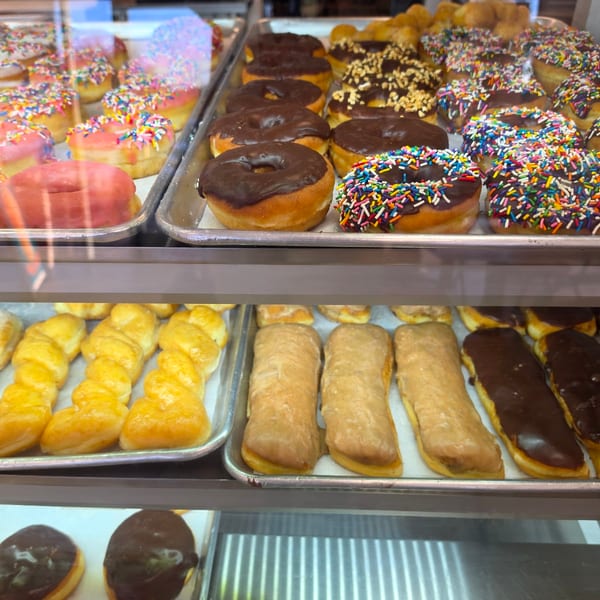 Variety of glazed, chocolate, and sprinkle donuts with other pastries in a bakery display case.